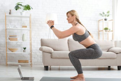 Woman with fitness tracker crouches and watches at laptop in living room interior